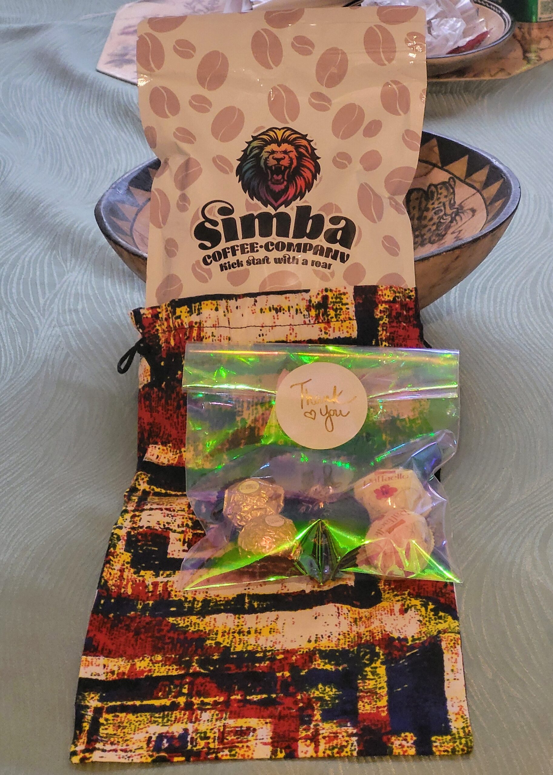 A colorful bag of Simba Coffee Company coffee sits next to a treat bag labeled "Thank You," containing chocolates and candies, on a patterned tablecloth.